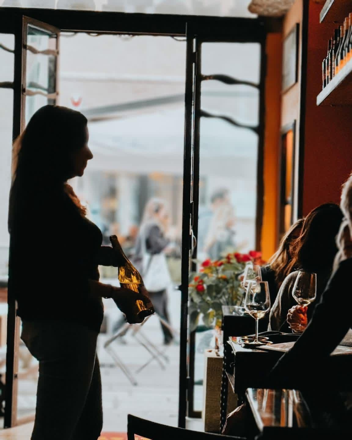 Una collaboratrice di Enotequa all'ingresso con una bottiglia di vino bianco, clienti in degustazione al tavolo