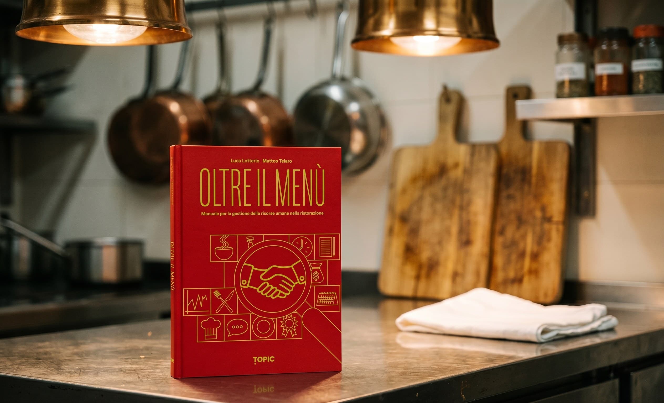 The book Oltre il Menù by Luca Lotterio and Matteo Telaro resting on the work surface of a professional kitchen, with copper pans hanging and cutting boards in the background